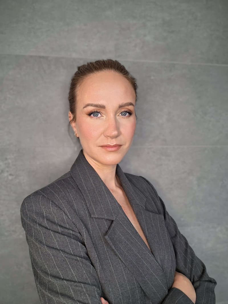 Tatjana Kadak wearing a dark pinstripe blazer, standing confidently with arms crossed in front of a gray background.